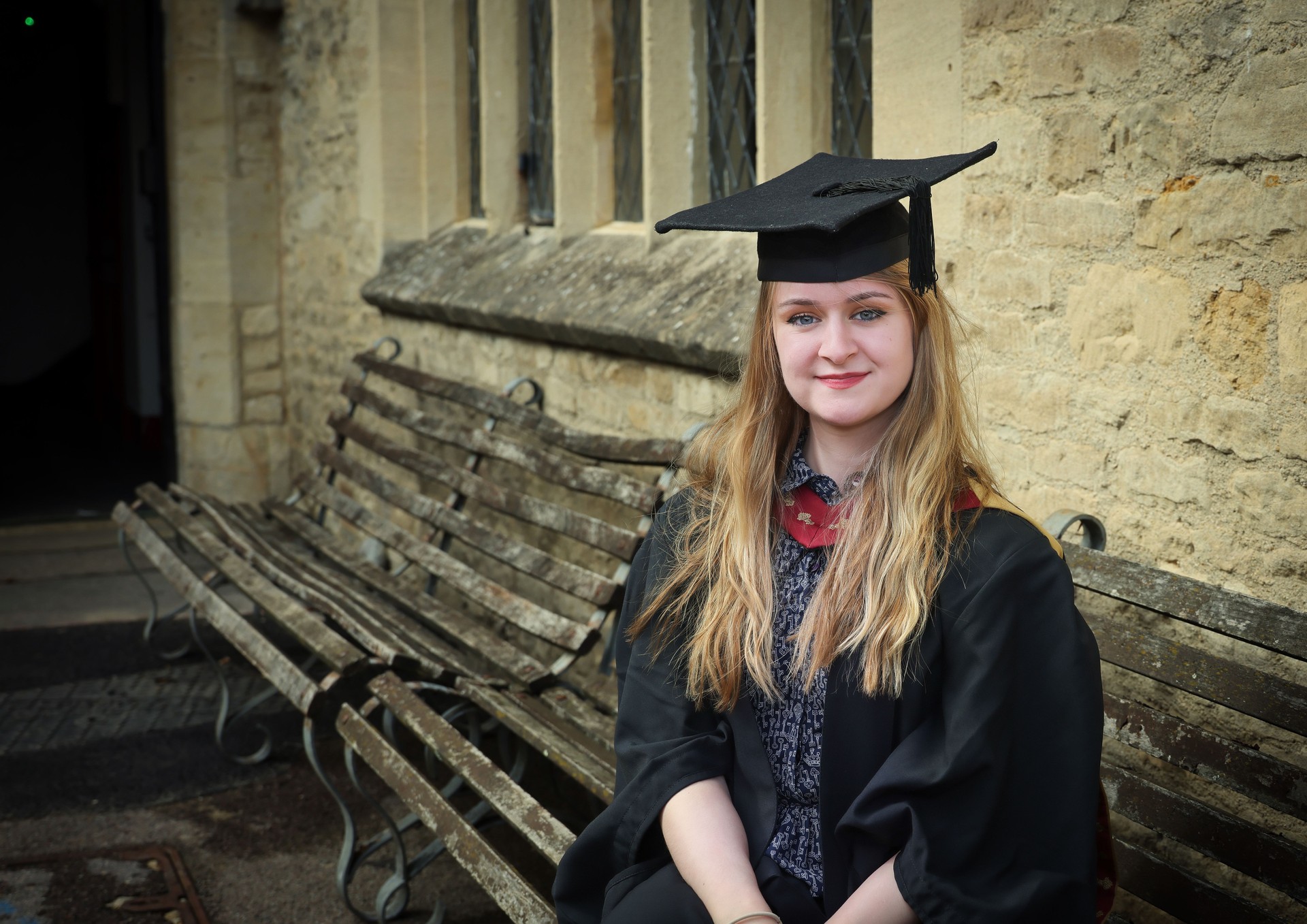 Proud, smiling and happy young woman smiling as she sits casually on an old bench on her Masters Graduation ceremony day.