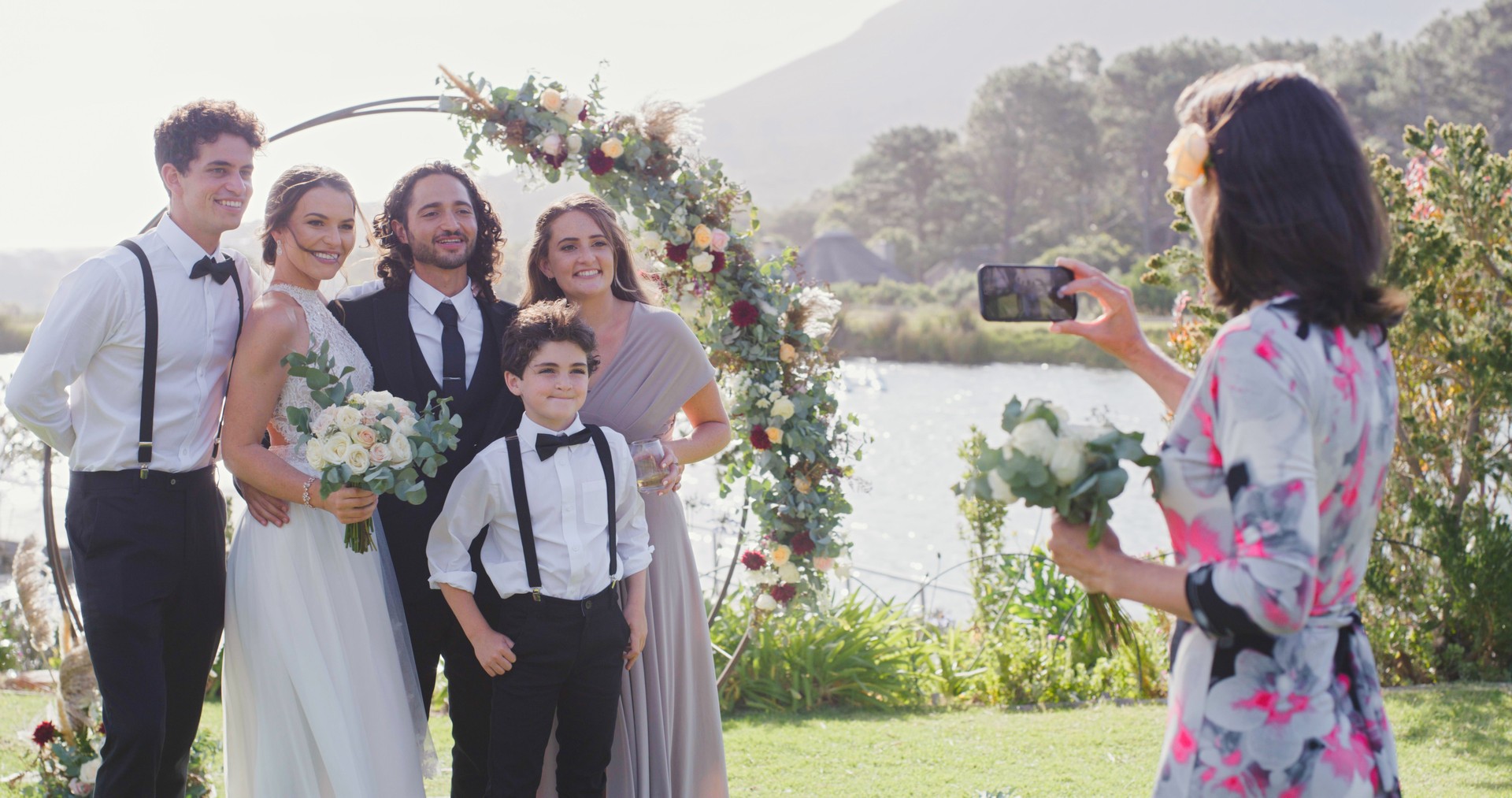 Family, couple and wedding portrait with flowers as photographer with a bouquet at a lake field nature event. Happy mother, children and bride smile with lovely groom in marriage celebration together